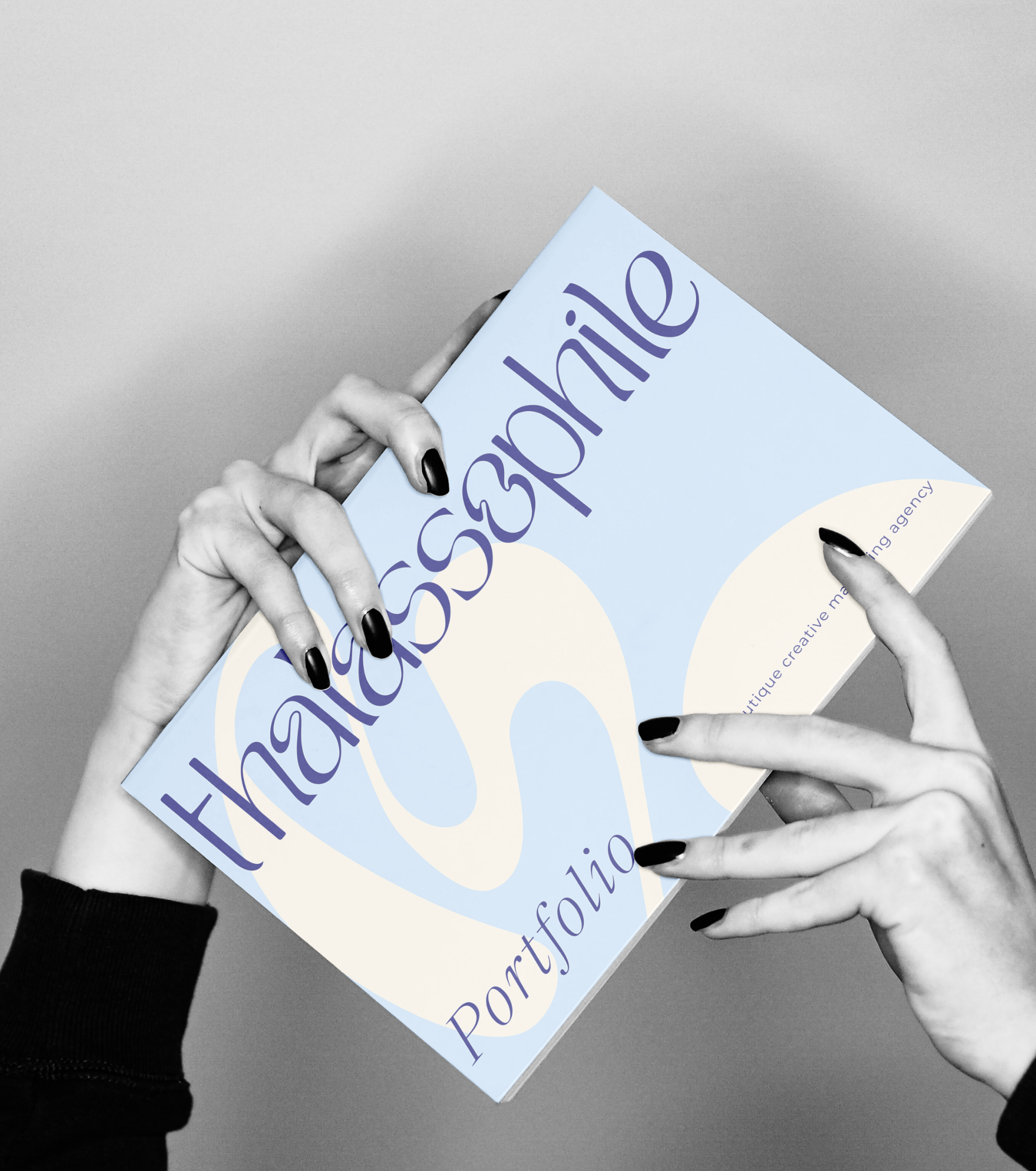 Hands holding the thalassophile studio portfolio in a stylish, minimalist setup. The portfolio cover features the thalassophile studio logo in blue and purple tones with the words ‘A boutique creative marketing agency.’ This image highlights thalassophile studio’s sophisticated branding approach, appealing to brands seeking expert marketing and visual storytelling services.