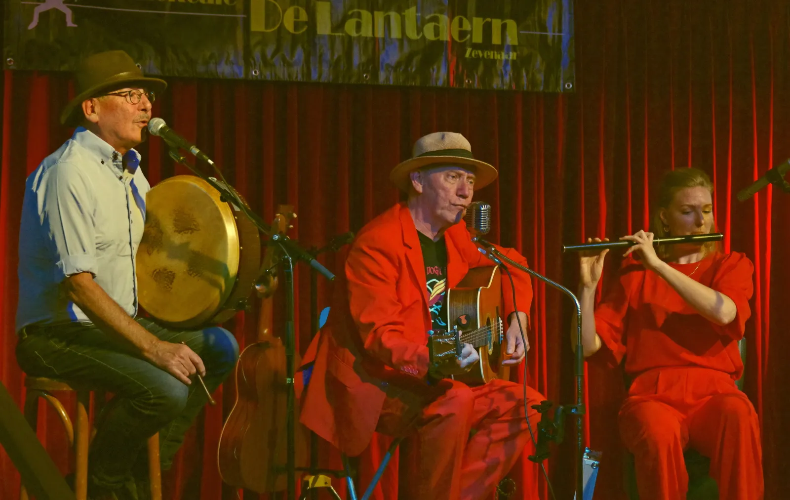 Zanger met bodhrán, gitarist in rood pak en fluitiste tijdens een concert in De Lantaern