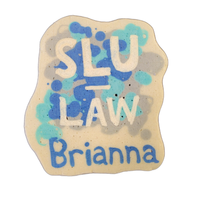Pancake art featuring the text 'SLU LAW' in large white block letters on the upper portion and the name 'Brianna' in blue handwritten-style letters on the lower portion, set against a cream-colored irregular blob background with blue and teal splatter shapes surrounding the central text. The piece…