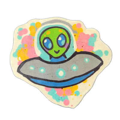 Pancake art depicting Trav, a round-headed green alien character riding in a gray flying saucer with a blue underside, set against a background of scattered pink, teal, and yellow circles and blobs on a light cream field. Bold dark outlines define the alien's large blue eyes, small smile, and the d…
