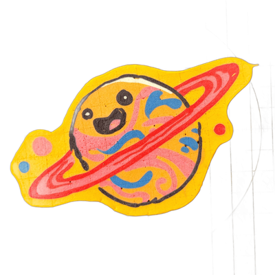 Pancake art depicting Saturnina (Nina), an original JoP character portrayed as a cartoon planet with Saturn-style rings encircling a round planetary body; the planet has a cartoon face with large black eye markings, an open mouth, and swirling surface patterns in pink and blue against an orange-yel…