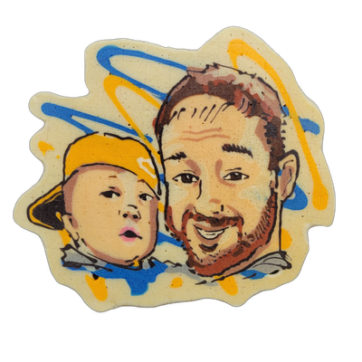 Pancake art depicting two faces side by side: a young child on the left wearing a yellow cap, with a slightly open mouth and wide eyes, and an adult on the right with short brown hair, a beard, and a broad smile. Rendered with bold dark outlines and warm skin-tone fill batter, accented by looping b…