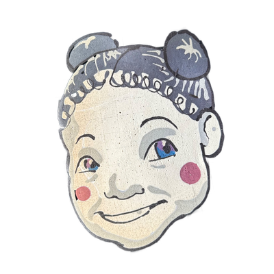 Pancake art portrait of a child's face with short, curly dark hair styled with two rounded bun-like shapes at the top of the head, large eyes with blue and purple irises, arched brows, a small nose, a slight closed-mouth smile, and circular pink blush marks on each cheek. Drawn by Wella with fine b…
