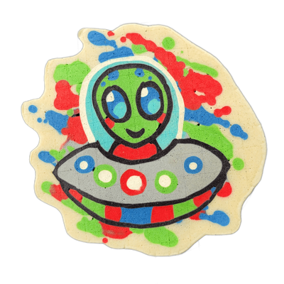 Pancake art depicting Trav, a green-skinned cartoon alien with large blue oval eyes and a small smile, shown from the chest up inside a light blue helmet or dome, seated in a gray flying saucer decorated with white-centered circles in red, green, and blue; a red scarf is visible at the neck area. T…