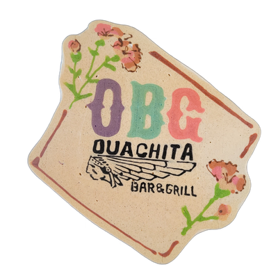Pancake art of the OBG (Ouachita Bar and Grill) logo, consisting of the large initials 'OBG' in purple, teal, and coral letters above the full name 'OUACHITA BAR & GRILL' in bold black text, with a stylized Native American headdress illustration centered between the name lines, a rust-colored decor…