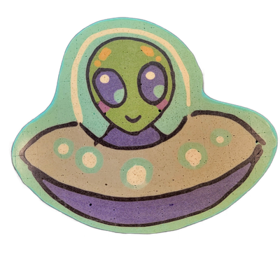Pancake art depicting Trav, a green-skinned alien character with large purple eyes, rosy cheeks, and orange spots on the top of the head, shown from the shoulders up inside a flying saucer with a domed cockpit area, a tan disc body decorated with teal-ringed circular markings, and a purple undercar…