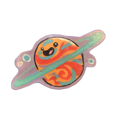 Saturnina, a JoP original character depicted as a cartoon planet with Saturn-style rings, shown as a sphere with a smiling face featuring large dark eyes and an open mouth, encircled by a diagonal ring band, with small star or moon shapes flanking the planet on either side. The pancake art uses bol…