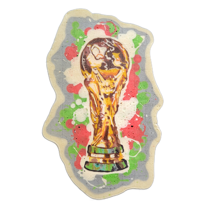 Pancake art depicting the FIFA World Cup trophy — a gold figurine of two human figures holding up a globe, set on a tiered base — surrounded by paint-splatter-style blobs in green, red, and white batter against a gray background. The trophy is rendered with bold dark outlines and layered warm gold…