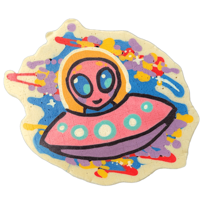 Pancake art depicting Trav, a cartoon alien character with a large oval head, oversized teal eyes, and a small smile, seated inside a pink flying saucer with teal circular porthole details and a purple underside, surrounded by a splattered background of blue, purple, yellow, orange, and red batter…