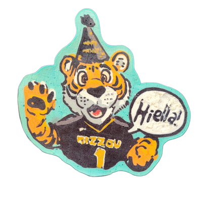 Pancake art depicting a cartoon tiger wearing a black jersey labeled 'Mizzou' and the number 1, with a black-and-gold party hat on its head, one paw raised in a wave, and a speech bubble to its right containing the text 'Hiella!'; the figure is set against a teal-colored blobby background shape. Th…