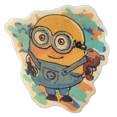 A round yellow cartoon character with two large circular goggles over both eyes, a wide mouth curved into a slight smile, wearing blue denim overalls, holding a small brown teddy bear in one hand, rendered in pancake art on a light cream base with irregular edges. Bold dark outlines define the figu…