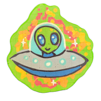 Pancake art depicting Trav, a JoP original character: a green-skinned alien with large blue oval eyes and a small smile, seated inside a gray flying saucer with teal circular lights along its rim, enclosed in a domed helmet or cockpit bubble rendered in light blue and white; the character wears a b…