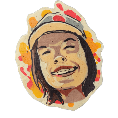 Pancake art portrait of a smiling person with shoulder-length dark hair, braces on their teeth, and a gray cap with red detailing on top of their head, rendered against a background of red, orange, and yellow batter splotches. The portrait uses bold dark outlines, a warm skin-tone palette of tans a…