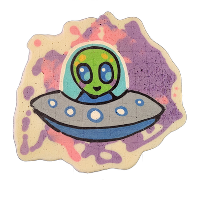 Pancake art depicting Trav, a green-skinned alien character with large blue eyes and yellow spots on his head, seated inside a gray and blue flying saucer with circular porthole markings along its rim, against a background of splattered violet and pink batter. Rendered by Dan with bold black outlin…
