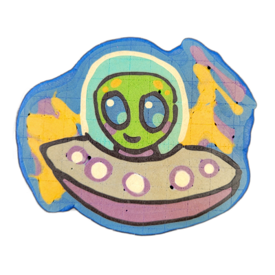 Pancake art depicting Trav, a green-skinned alien character with large dark oval eyes, orange spots on the forehead, and a small smile, seated inside a domed flying saucer cockpit; a purple scarf is visible at the neck, and the saucer's underside features a row of circular porthole-like lights in w…