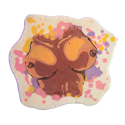 Pancake art depicting a pair of large, dark-toned breasts with nipples, rendered in brown and orange batter tones against a white background with scattered paint-splatter-style dots in pink, yellow, and violet. Bold black outlines define the forms, with orange highlight areas contrasting against da…