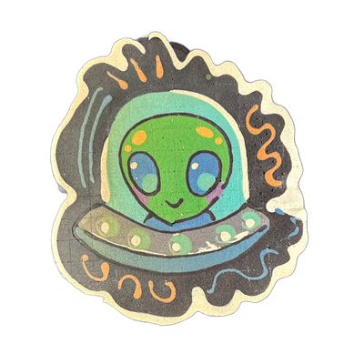 Pancake art depicting Trav, a green alien character with large blue eyes, yellow-orange spots on his head, and a small smiling mouth with pink cheeks, seated inside a gray flying saucer with circular markings along its rim, under a teal-tinted dome helmet; drawn by Wella in approximately 9 minutes…