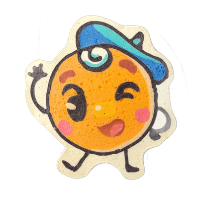 Pancake art depicting Jacques Floppes, a round soufflé-pancake character with a cartoon face featuring one wide open eye and one closed eye, pink circular cheeks, a small open mouth, and chocolate-chip-style spots on an orange-toned body, wearing a blue and teal cap tilted to one side with a teal s…