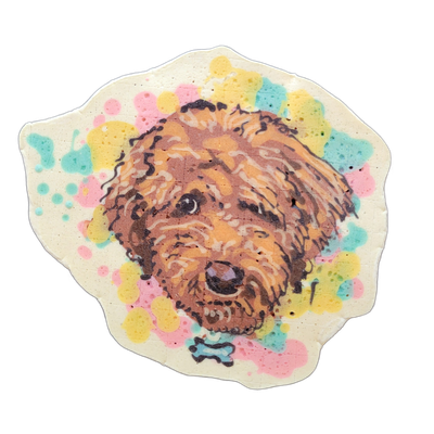 Pancake art portrait of a dog named Teddy, shown from the neck up facing forward, with long wavy golden-brown fur, dark eyes, a rounded muzzle, and a small collar tag shaped like a bone rendered in teal batter at the bottom center. The portrait uses fine dark outlines with thin white highlight line…