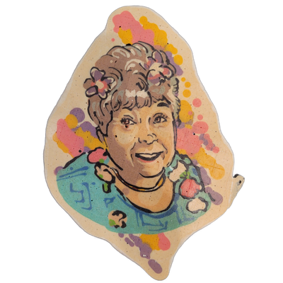 Pancake art portrait of a smiling woman with short, curly gray-brown hair adorned with small flower shapes, wearing a teal top with a floral pattern and a dark necklace, shown from roughly the shoulders up against a background of splattered shapes in violet, pink, and yellow. Dan rendered the piece…