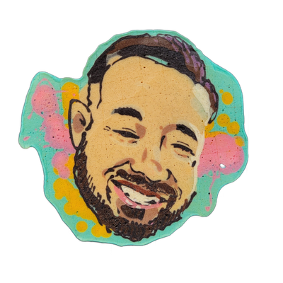 Pancake art portrait of a person named Erin, showing a smiling face with short dark braided or twisted hair, a beard and mustache, dark eyebrows, and squinting eyes mid-laugh, with ears visible on both sides. The face is rendered in warm tan and brown tones with bold dark brown outlines, set agains…