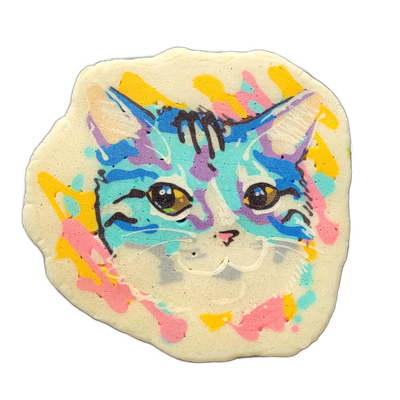 A cat face drawn in pancake art, centered and filling most of the composition, with pointed ears, amber-colored eyes, a small pink nose, and white whisker lines radiating from the muzzle area. The face is rendered with bold black outlines and filled with patches of teal, blue, violet, and white bat…