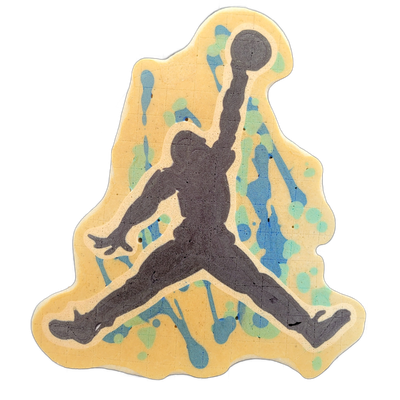 Pancake art depicting the silhouette figure from the Air Jordan 'Jumpman' logo — a human figure in mid-air with one arm raised holding a ball overhead and legs split wide apart, rendered in dark gray-brown batter against a yellow-cream background with blue and teal paint-splatter-style shapes scatt…