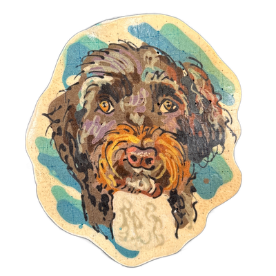 Pancake art portrait of a dog named Chester, depicted face-on with a broad muzzle, amber eyes, a pink nose, and long wavy fur rendered in brown, black, tan, and orange tones against a teal and blue background with a cream border. The piece uses bold black outlines and dense layered batter strokes t…