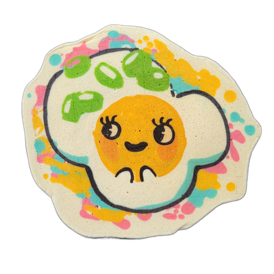 Pancake art depicting Sunny, an original JoP character designed as a fried egg with a cartoon face: a round orange yolk as her head, with drawn-on eyelashes, dot eyes with small highlights, rosy cheeks, and a small smile, set against an irregular white egg-white shape outlined in bold black lines,…
