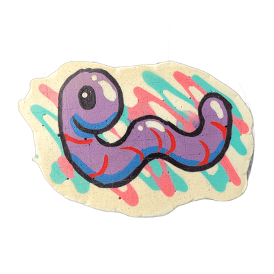 Pancake art depicting Hermie the Wurmy, an original JoP character rendered as a purple worm-like figure with a large circular head featuring a single eye, a curved mouth with blue marks, and a sinuous body that tapers into a curled tail, all set against a cream-colored irregular background decorate…