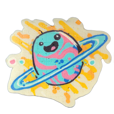 Saturnina, an original JoP character depicted as a round cartoon planet with two small eyes, an open smiling mouth with a pink tongue, and wavy pink bands across a teal body, encircled by a blue and white Saturn-style ring. The pancake art is drawn by Dan with bold black outlines, filled in teal, p…