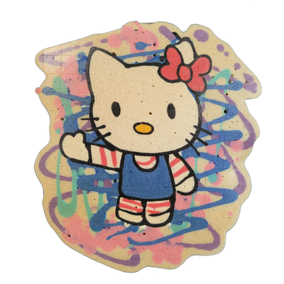A pancake art depiction of Hello Kitty, the Sanrio character, shown in a front-facing standing pose with one arm raised in a wave, wearing a blue dress over a pink-and-white striped shirt, striped shoes, and a red bow on the upper left side of her head, with a white oval face, small black oval eyes…