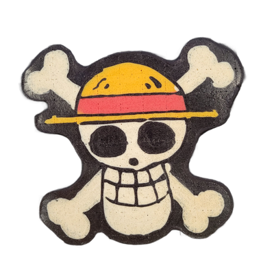 Pancake art of a skull-and-crossbones design wearing a straw hat with a red band, rendered as a flat cutout shape against a dark background; the skull has large round eye sockets, a wide grin with rectangular teeth, and a yellow brim line extending horizontally across the face, with two pairs of cr…