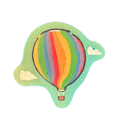 A hot air balloon with vertical rainbow-colored stripes — green, yellow, orange, red, blue, and purple — floats against a teal sky background with two small white clouds, one on each side; a small basket hangs beneath the balloon with a red burner detail visible. Drawn by Wella in pancake art using…