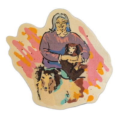 Pancake art depicting a person with gray hair holding a small brown dog in their arms, with a larger brown-and-tan long-haired dog positioned in the lower left foreground. The piece uses bold black outlines with fill areas in purple, brown, blue, and teal, against a cream-colored background splashe…
