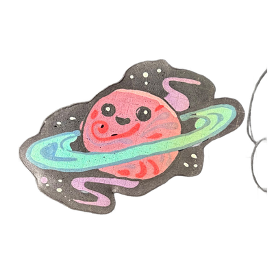 Pancake art of Saturnina (Nina), a JoP original character depicted as a round, cartoonish planet with a smiling face, large round eyes, rosy cheeks, and a small open mouth, encircled by a broad ring rendered in teal, green, and blue gradients, with a secondary curved purple-pink element and a dark…