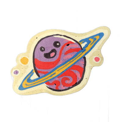 Pancake art depicting Saturnina (Nina), an original JoP character portrayed as a cartoon planet with Saturn-style rings and a cartoon face featuring large oval eyes and an open smiling mouth. Bold black outlines define the form; the planet body is filled with swirling red and violet batter, the rin…