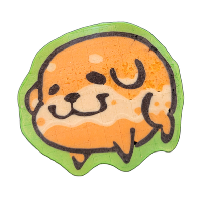 Pancake art depicting Poffridge, a Shiba Inu-shaped sentient poffertje from the JoP cast, shown as a round, compact head-and-body form with small dark facial features — a round nose, curved mouth, dot eyes, and two raised paw-like shapes visible at the lower right — set against a dripping green bor…