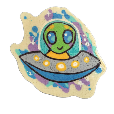 Pancake art of Trav, a green-skinned alien character with large blue oval eyes and a small smile, seated inside a gray flying saucer with yellow circular lights along its rim, wearing an orange collar or neck piece, set against a background of splattered blue, teal, and purple shapes with small whi…