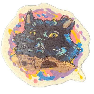 A black cat's face fills most of the composition, shown from roughly the shoulders up, with yellow-green eyes, visible whiskers, and ears with tufted fur; the cat appears to be resting its chin on a flat surface rendered in tan and brown tones. The pancake art uses bold dark outlines for the cat's…