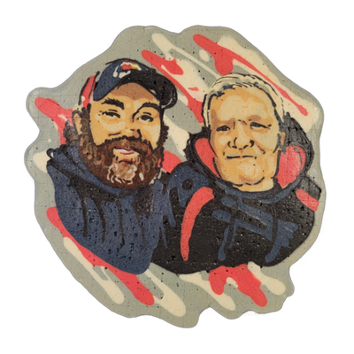 Pancake art portrait of two men shown from the shoulders up, side by side: the man on the left has a full dark beard, a dark baseball cap with a small logo, and wears a dark hoodie with red and navy detailing; the man on the right has short white or light gray hair, wears headphones or ear protecti…