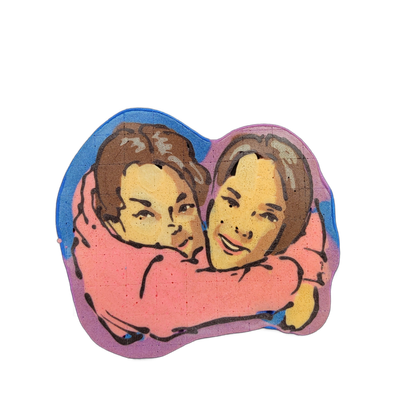 Pancake art depicting two people, labeled Millie and Lina, shown from roughly the shoulders up, with one person's arms wrapped around the other in an embrace; both have brown hair, yellow-toned skin rendered in batter, and dark brow and facial feature outlines. The piece uses bold black outlines ov…