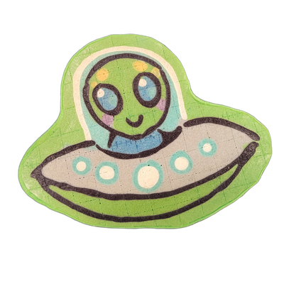 Pancake art depicting Trav, a green-skinned alien character with large blue eyes, small pink cheek marks, and yellow spots on the top of the head, seated inside a gray flying saucer with a transparent domed cockpit and three teal-rimmed white lights along the saucer's rim. Rendered by Dan with bold…