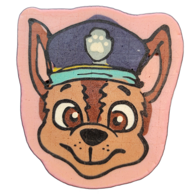 Pancake art depicting a cartoon dog face wearing a blue police-style cap with a paw print badge on the front, a teal brim, and a yellow band; the dog has large round brown eyes with white highlights, a black nose, and a smiling muzzle, with brown fur markings and two jagged dark stripes running ver…