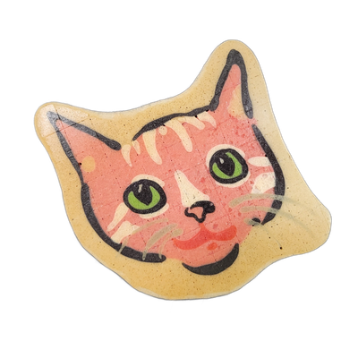A cat face rendered in pancake art, showing a front-facing view with pointed ears, green irises with black pupils, a small pink nose, and light-colored stripe markings across the forehead and cheeks against a pink-toned face, all set within a cream-colored border that follows the shape of the head.…