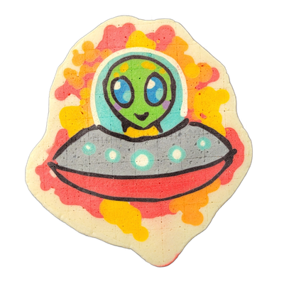 Pancake art of Trav, a green-skinned cartoon alien with large blue eyes, a small smile, and a light purple mark on the chin, wearing a transparent dome helmet with a yellow collar, piloting a gray flying saucer with teal and white circular lights along its rim, set against a background of red, oran…