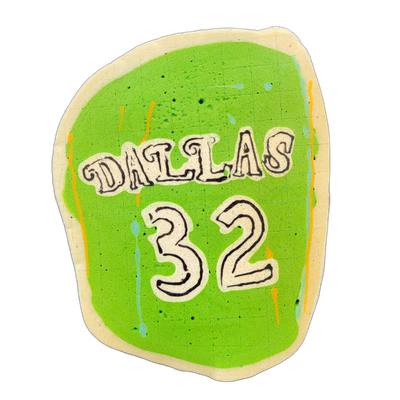 A pancake art piece shaped like a rounded rectangle depicting a sports jersey-style graphic with the word 'DALLAS' arched across the upper portion and the number '32' large below it, both rendered in cream-colored block lettering with black outlines on a bright lime-green background. The lettering…