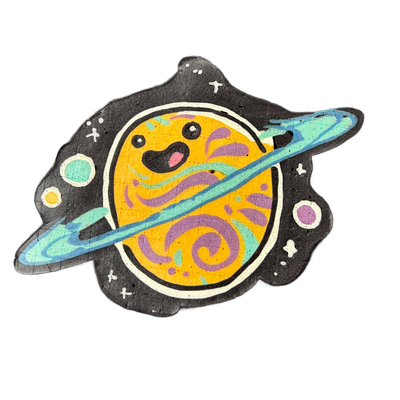 Saturnina, a JoP original character depicted as a round planet with a cartoon face — two solid black circular eyes, a small open mouth with a pink tongue or interior, and pink cheek marks — encircled by a tilted ring rendered in teal and blue gradients, set against a black irregular background shap…