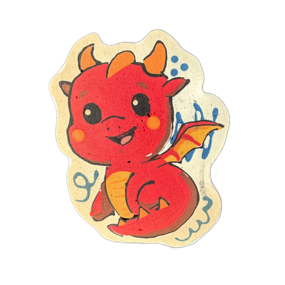 A small, round-bodied cartoon dragon rendered in pancake art, shown from the front in a seated pose with two small horns, large round eyes, rosy circular cheek markings, an open smiling mouth, small visible wings on its back, and a curled tail; the body is red with orange and yellow-orange accent a…