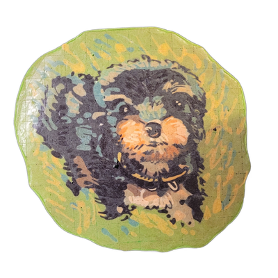 Pancake art of a small, dark-furred dog named Chemin, shown from the front in a seated or crouched pose, with tan and orange coloring on the muzzle and cheeks, a small black nose, dark eyes with white highlight dots, a visible tongue, and a yellow collar; the dog is surrounded by a green background…